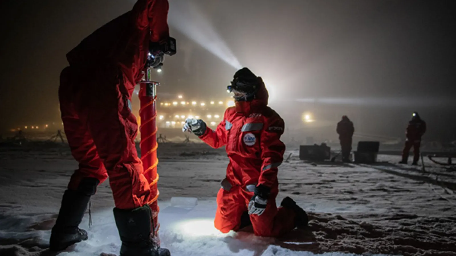 MOSAiC Ice Expedition team members drilling ice in the night. They are wearing bright red snow suits