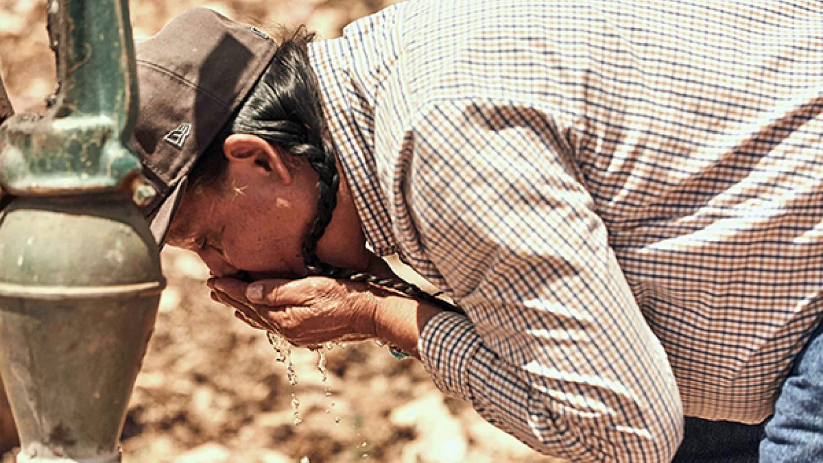 A person bends down to drink water from a water pump on this very dry land 