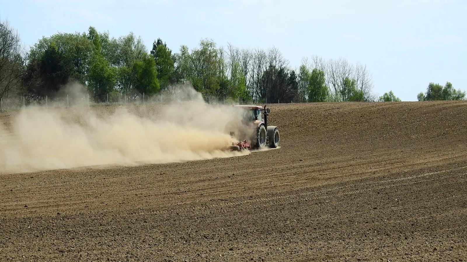 A tractor drives across barren agricultural land and kicks up dust behind it 