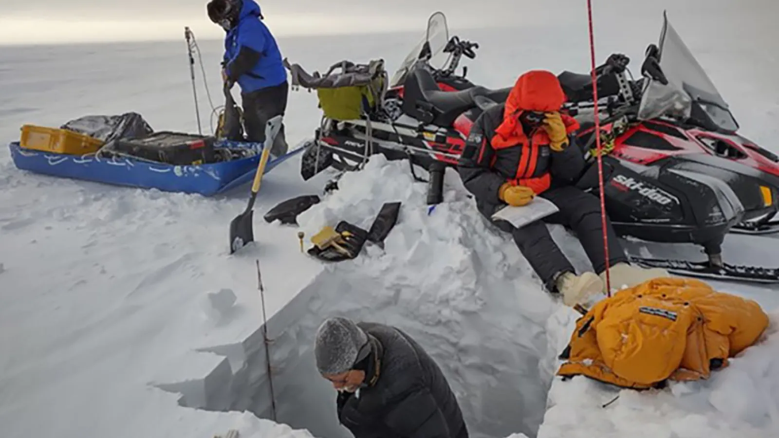 A man digs a snowpit in a snow field while another sits on snow pile next to snowmobiles writing in a notebook and another is working on equipment on a sled