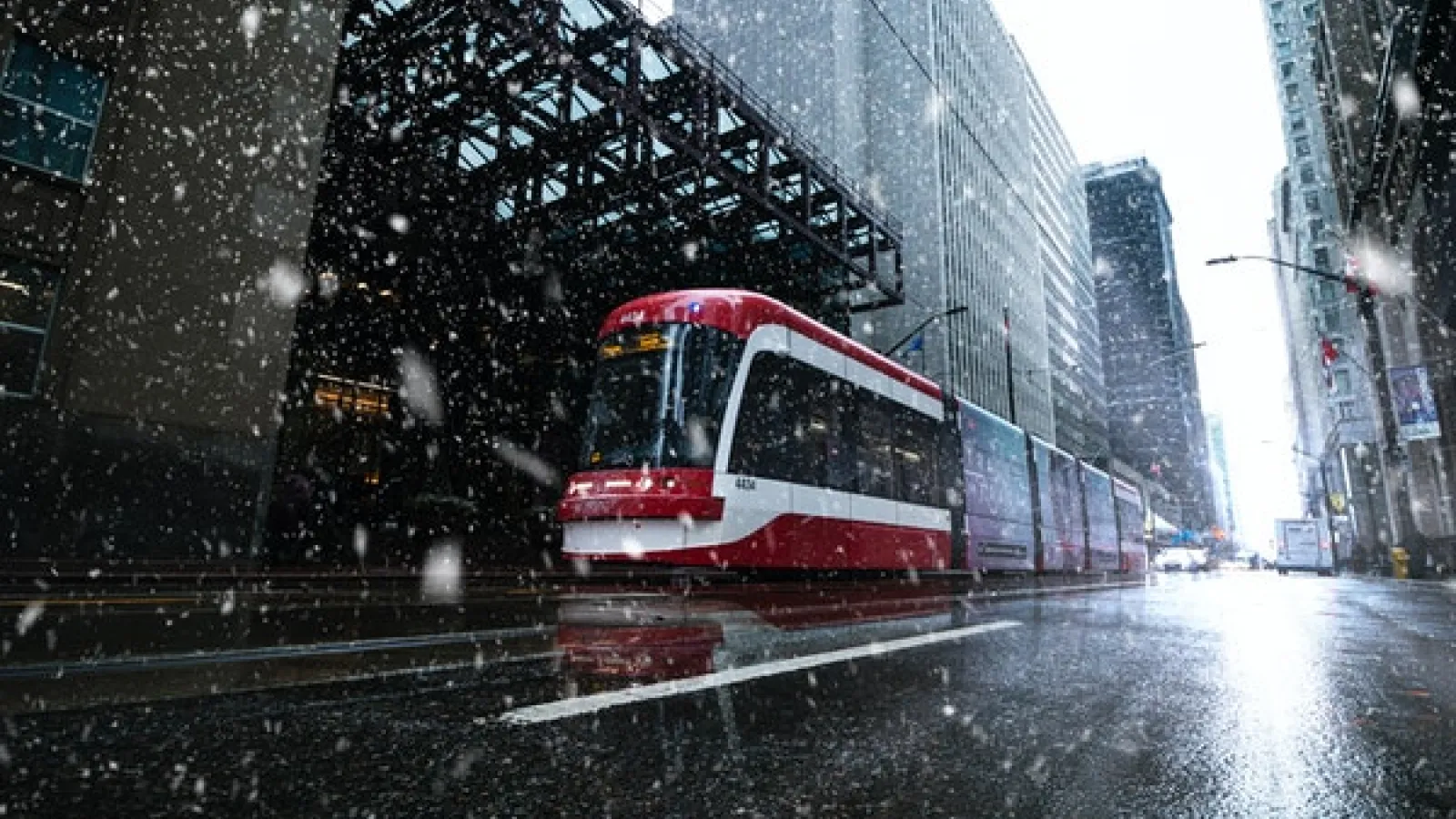 A red streetcar on a city street in the snow surrounded by tall and short buildings.