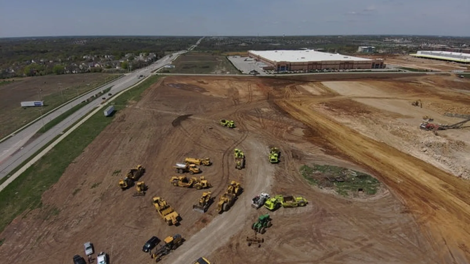 Sky view of freeway, construction site, equipment, vehicles, building and houses at a distance.