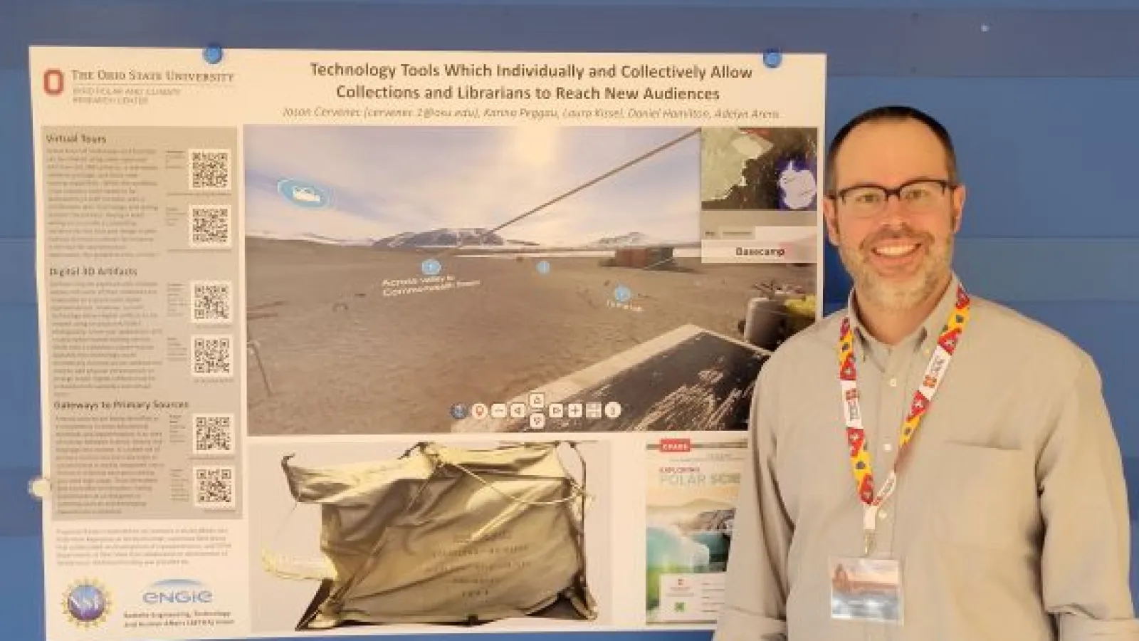 Jason with glasses in tan shirt with colorful lanyard standing to left of poster on blue wall