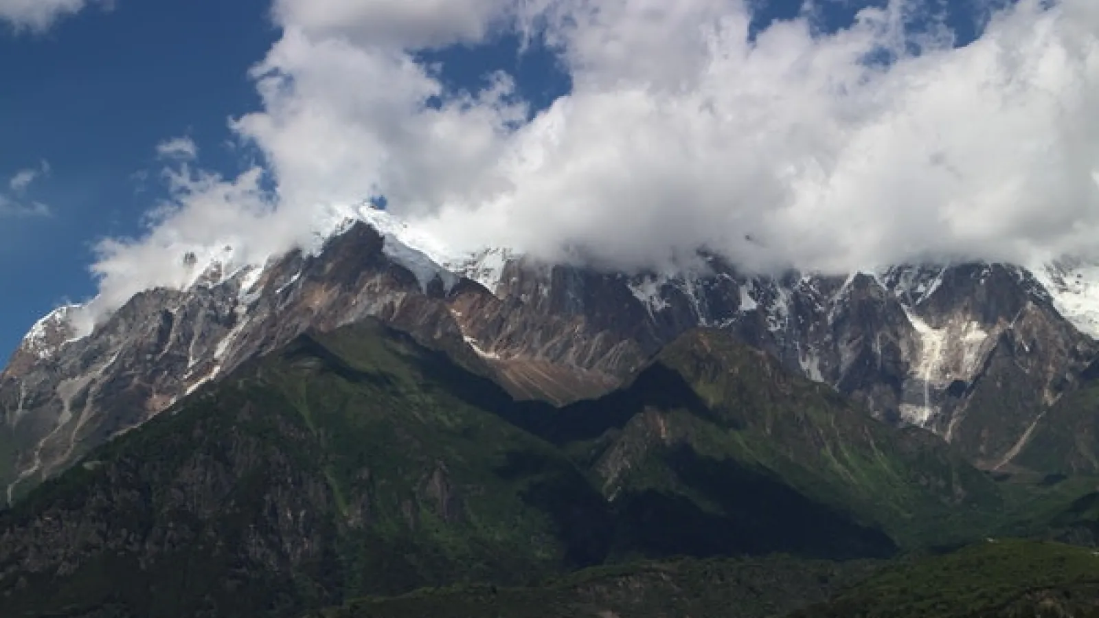 Snow-capped mountain range with trees at foothills and white clouds in a blue sky.