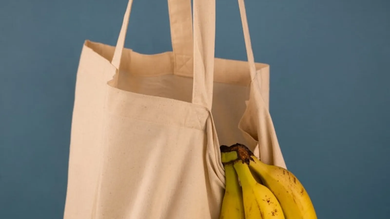 a hanging tan fabric shopping bag with a banana bunch hanging out with a gray-blue background