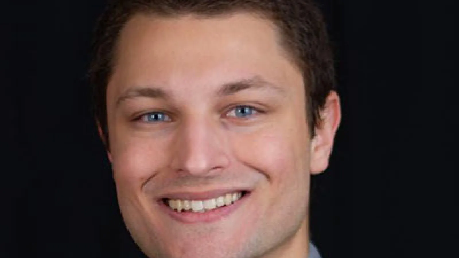 Portrait image of Austin Weber smiling wearing dark gray suit and tie with light gray collar shirt and black background.