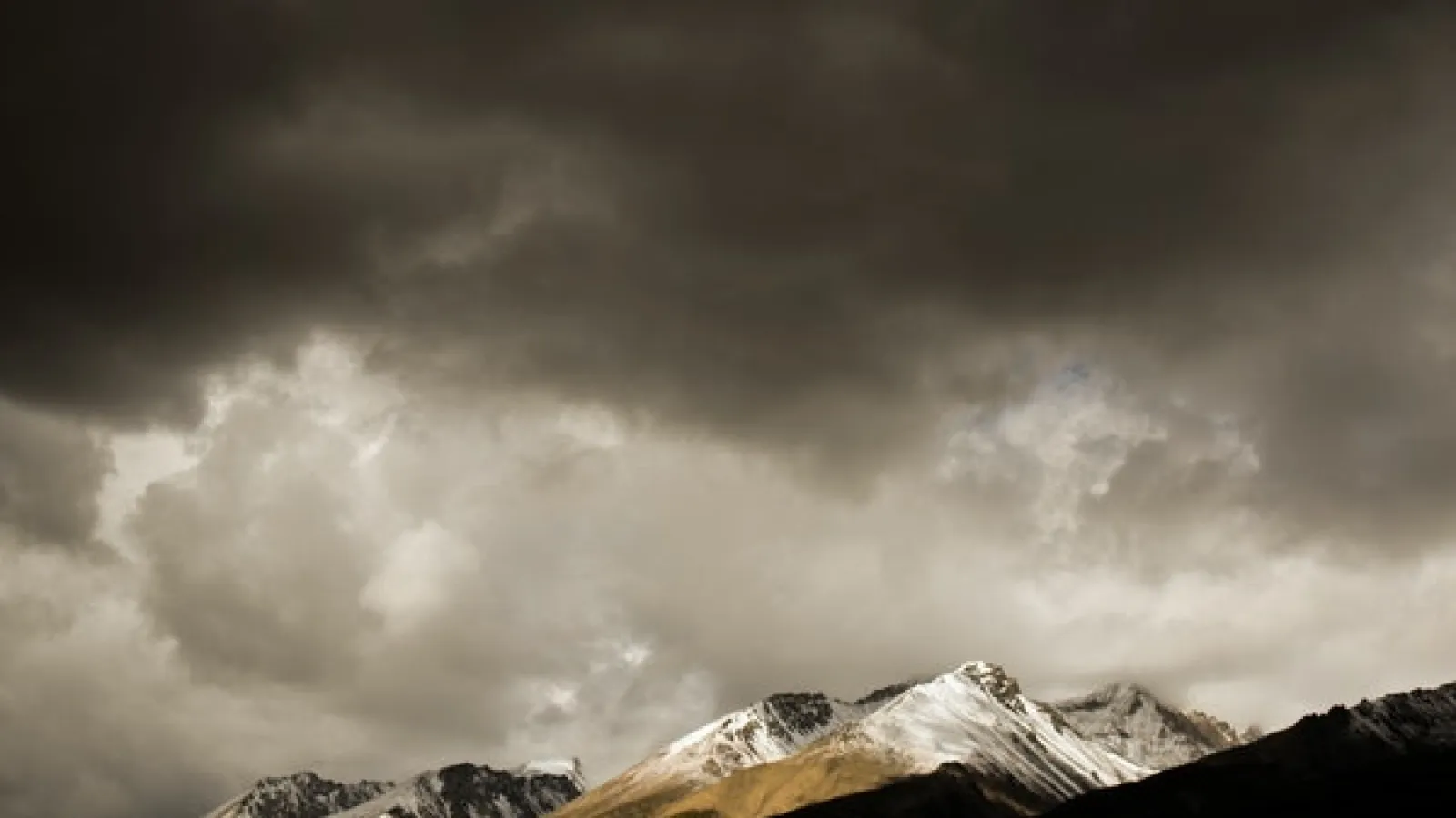 Snow covered, some treed and some brown mountains at a distance with dark cloud at the top but white clouds right about the mountains and dark mountains in the foreground.