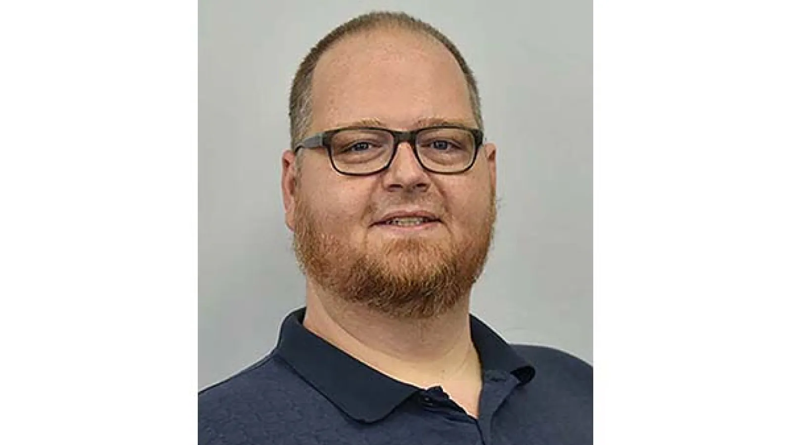 portrait of Aaron Wilson with light background wearing dark blue collar shirt and glassed with beard and short hair