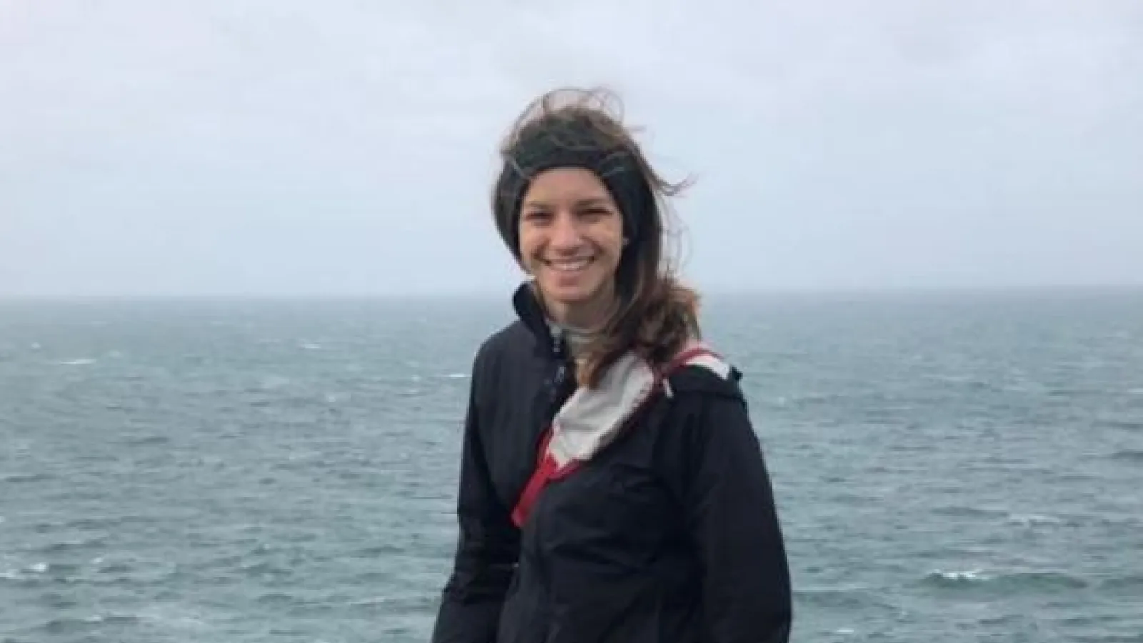 Devin Smith taking facing the camera and smiling while step onto brown rocks in front of body of water with light wave wearing blue jeans and dark jacket and head band with longer hair pulled back and blue skies with clouds