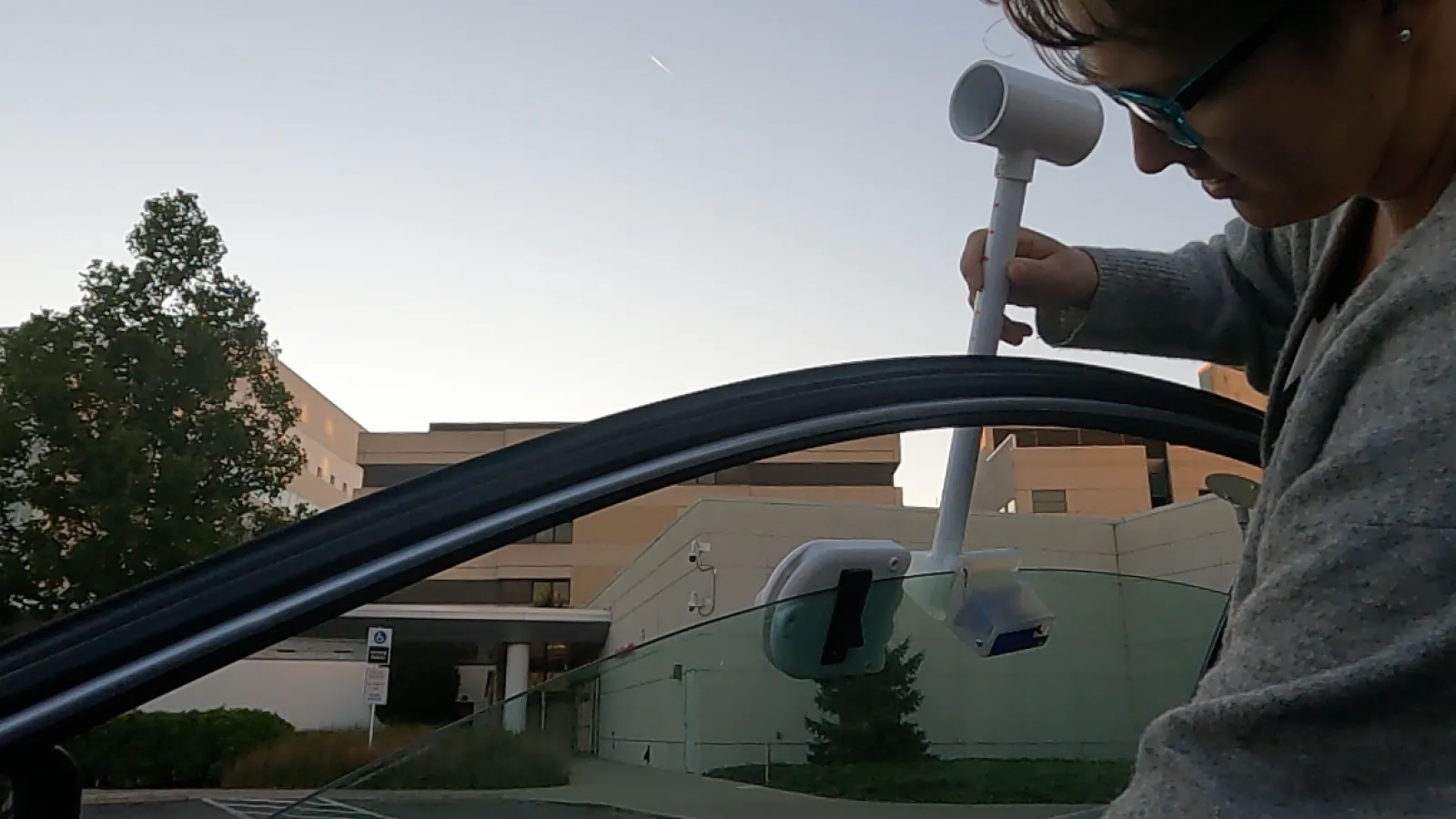 left side of Karina Peggau standing sideways wearing a gray sweater, glasses and pearl earing on right side of image with an open car door, three quarter open window installing  an apparatus on the window in a parking lot with a building and side walk and grass and trees in background