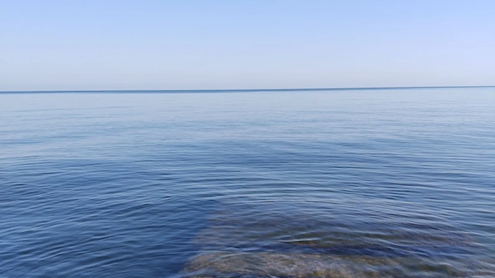 calm lake during the day with blue water and sky and silhouette of slabs of rock covered moss in forefront under water