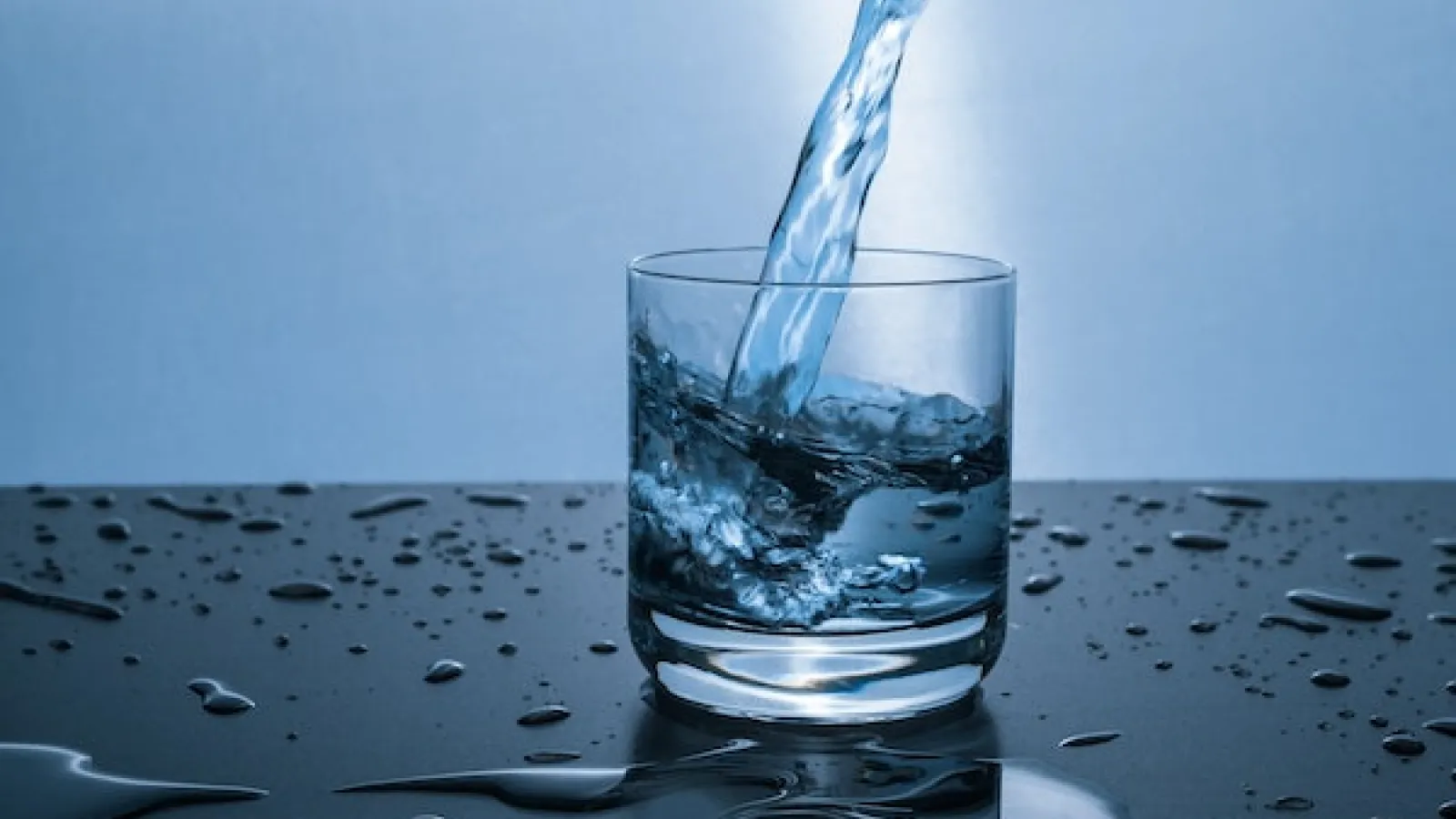 Clear water being poured from above into a short glass on a black shiny surface with water puddles and drops on it and a light blue background