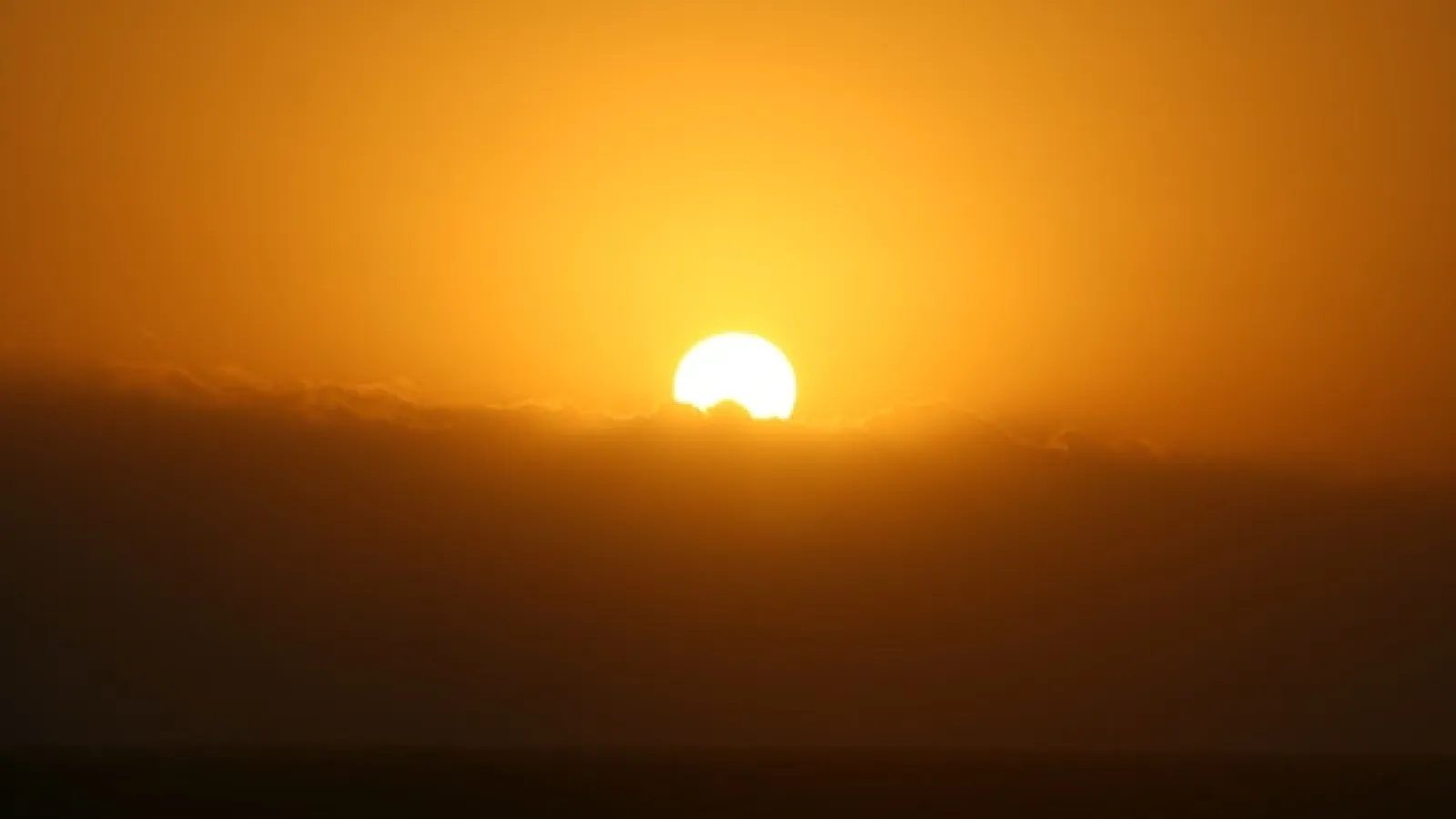An orange sun partially behind puffy clouds with the whole image as orange, with the top half orange sky and the bottom half clouds and what seems to be land at the bottom as a darker shade of orangish brown.