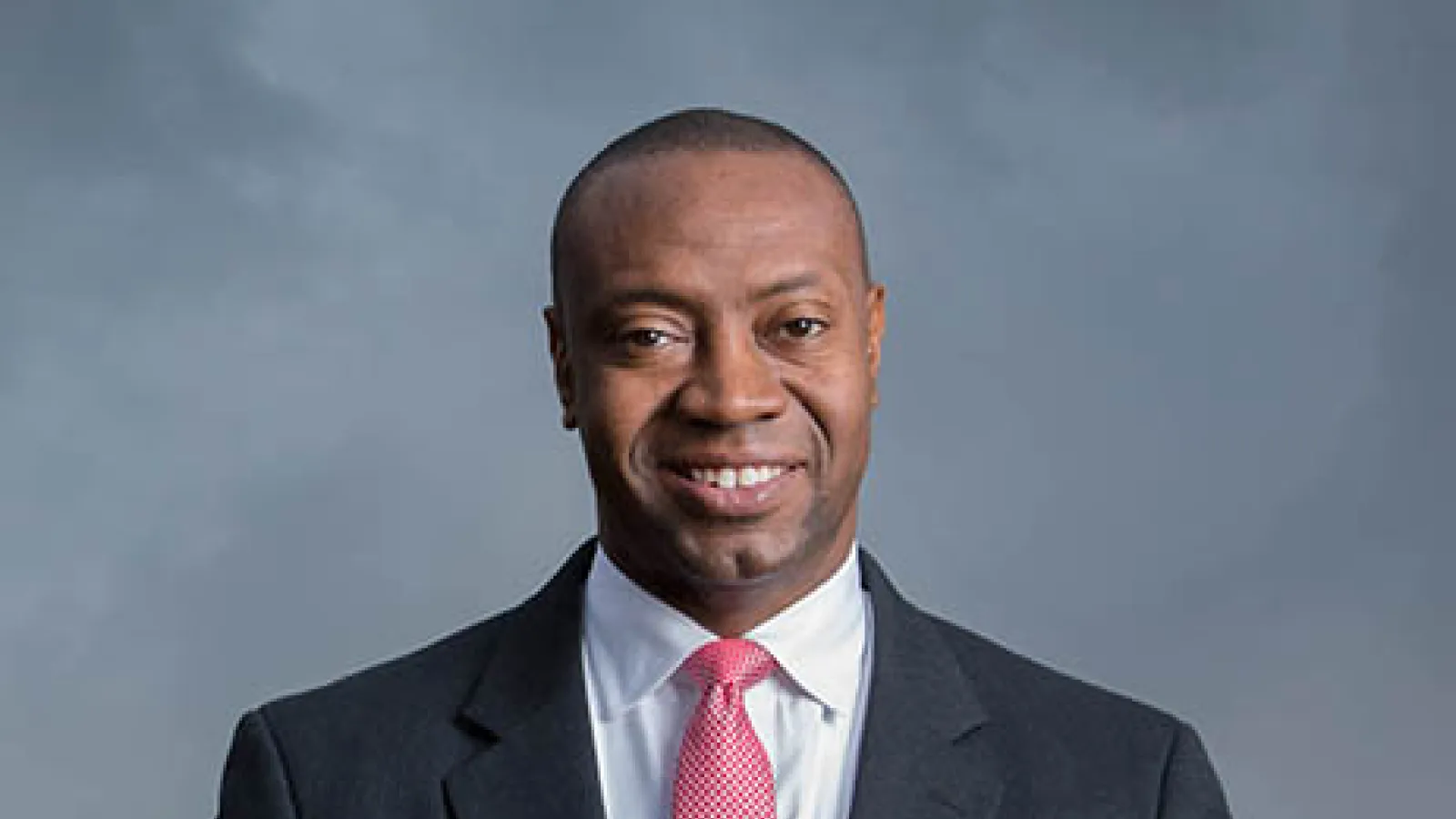 Portrait of Dr. J Marshall Shepherd with gray background smiling with very short hair wearing a white collar shirt and dark gray suit and pink rose tie and top and bottom of image have rose color