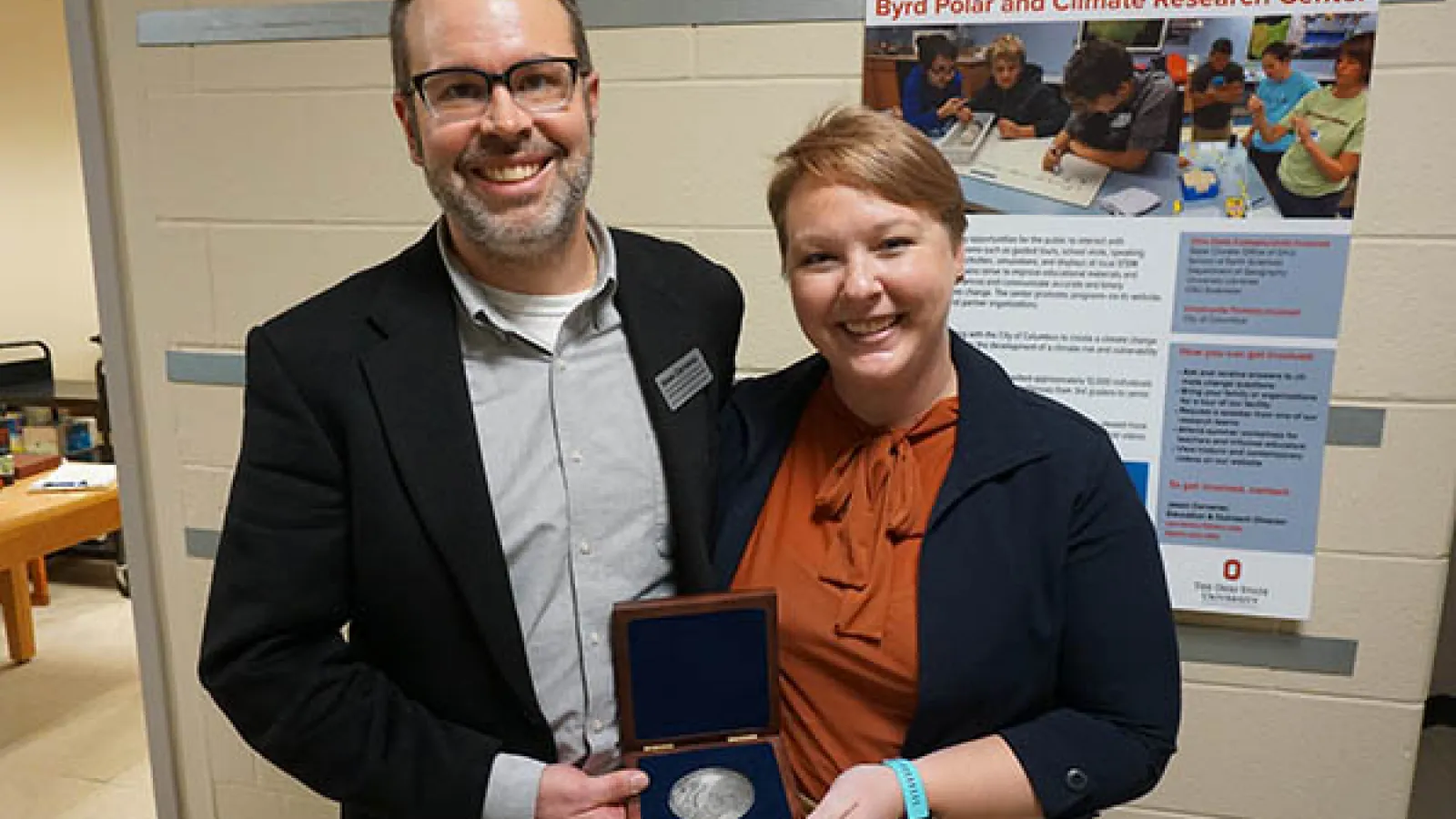 Jason Cervenec standing on the left side in the middle of the screen facing the camera with his right hand holding an open box with a round medal in it wearing a while collar shirt and a black blazer with glasses smiling while next to him Allison Chartrand is to his left holding that same box with her left hand wearing a black blazer and terracotta color silk shirt, light short hair and smiling