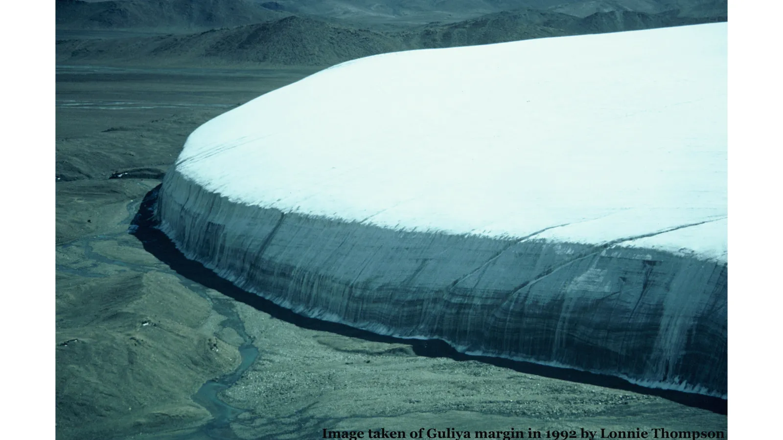 Blue-ish hue landscape and a massive wall of Guliya margin snow covered ice terminating to a plateau above the landscape dominating the image in the middle and the right of the photo 