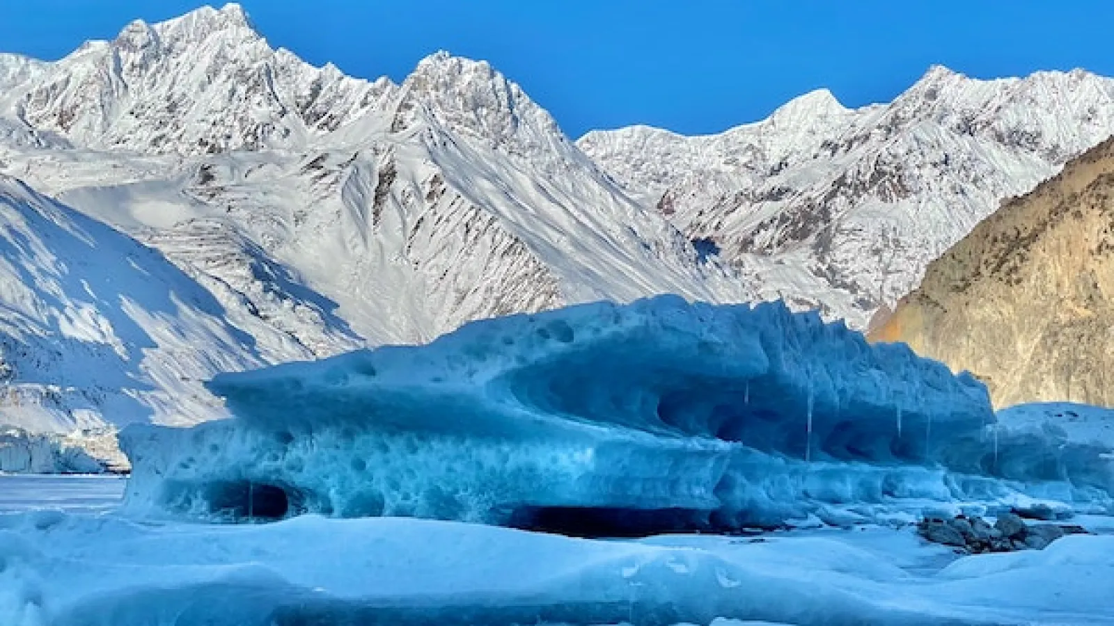 Snow covered mountain view with blue skies and ice in foreground and a dirt mountain on right side