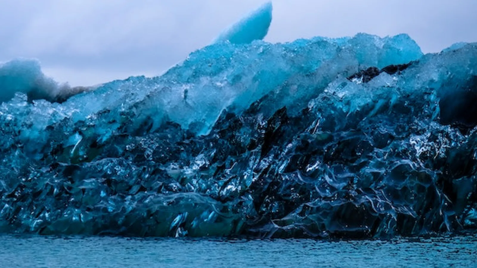 Large pieces of melting light blue and clear ice with jagged edges on black rocks next to a body of water with grey skies in the background  