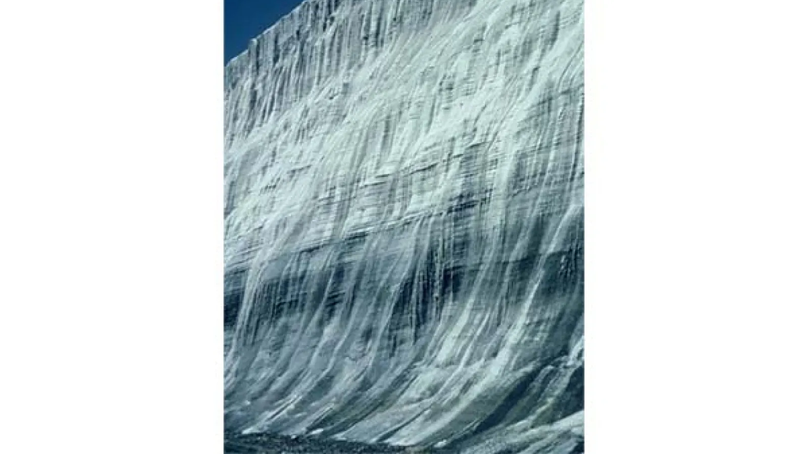 small image of a wall of ice with different layers hues of dark color with a small area of deep blue sky .