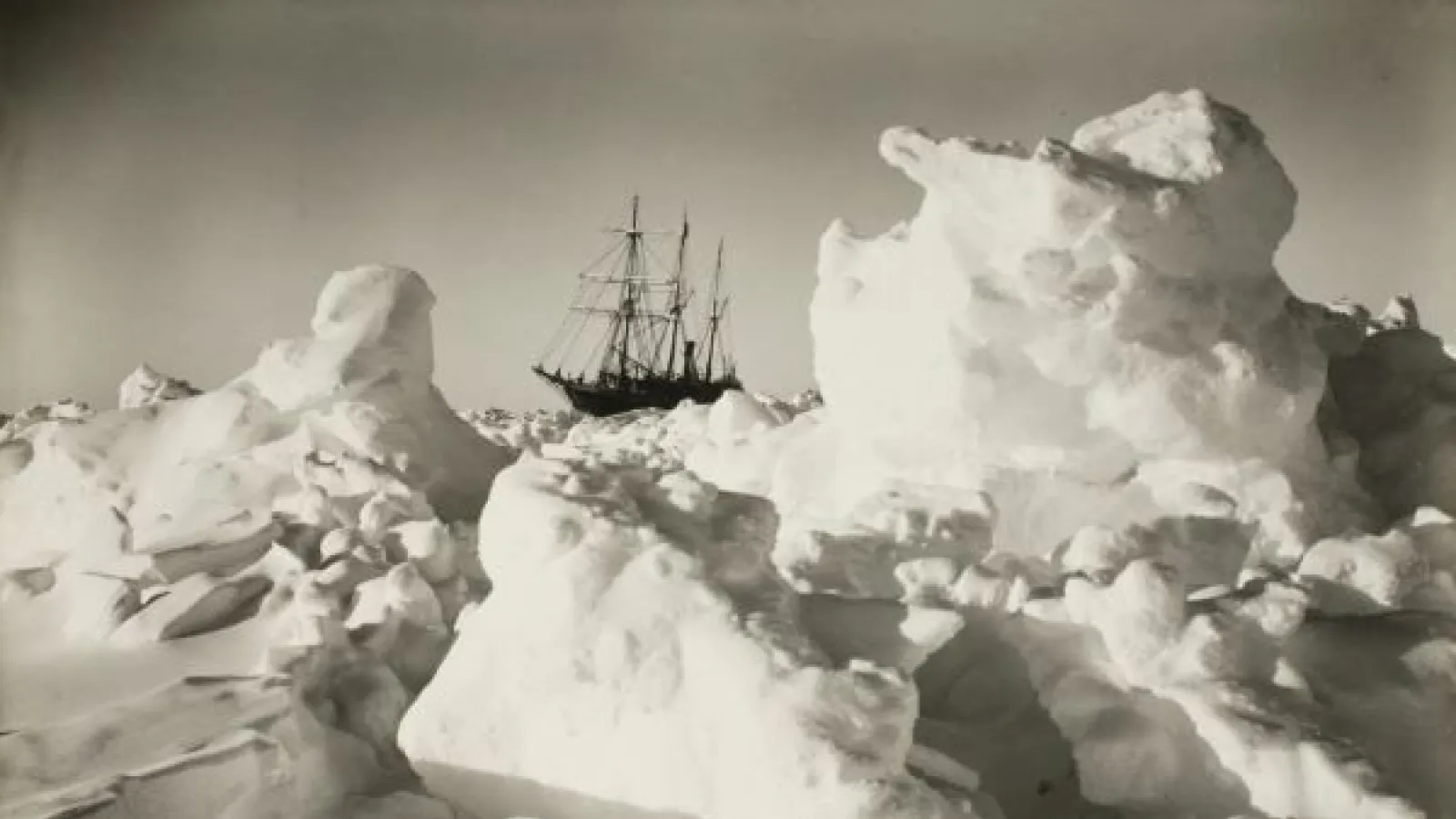 icebergs and crumpled ice in foreground and small ship in background. photo color is sepia. 