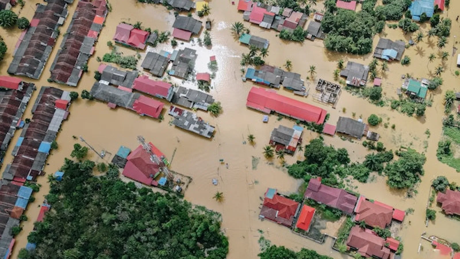 aerial image of flooded city with muddy waters covering roads and engulfing greenery and trees and building structures with blue and pink slanted roofs