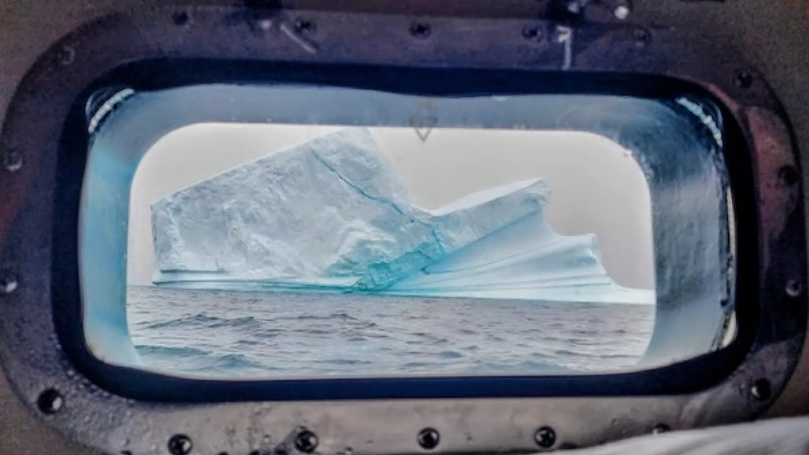 White sky and a blue white iceberg in the calm light blue water filling the porthole of a ship we look through with an elongated oval shape frame and rivets around the metal  frame