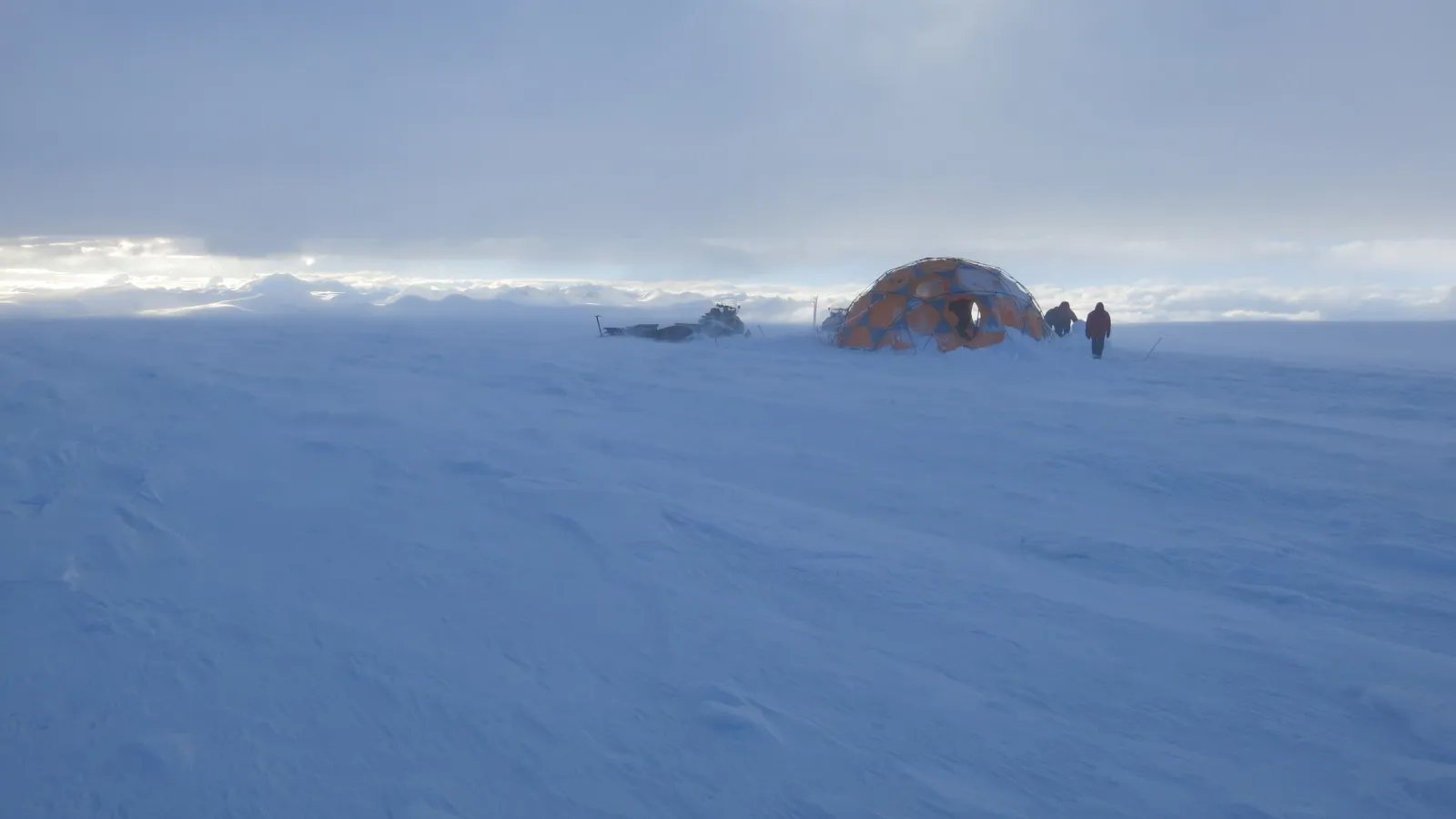 A tent in a distance on snow covered ground under sunny blue skies.
