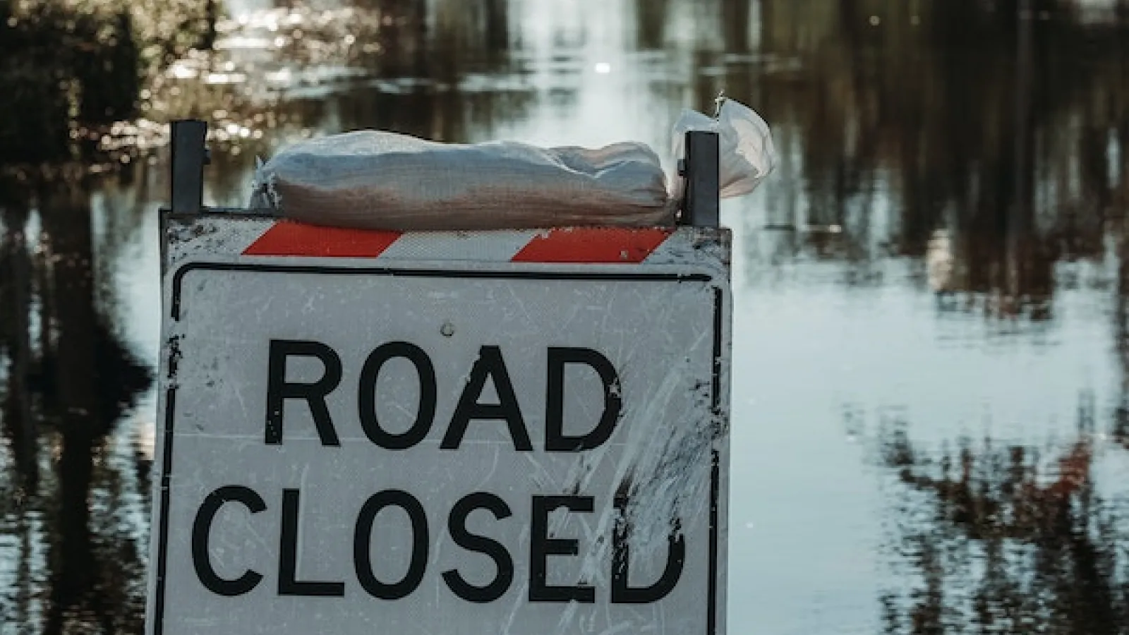 A Road Closed sign with a sandbag on it blocking a flooded road.