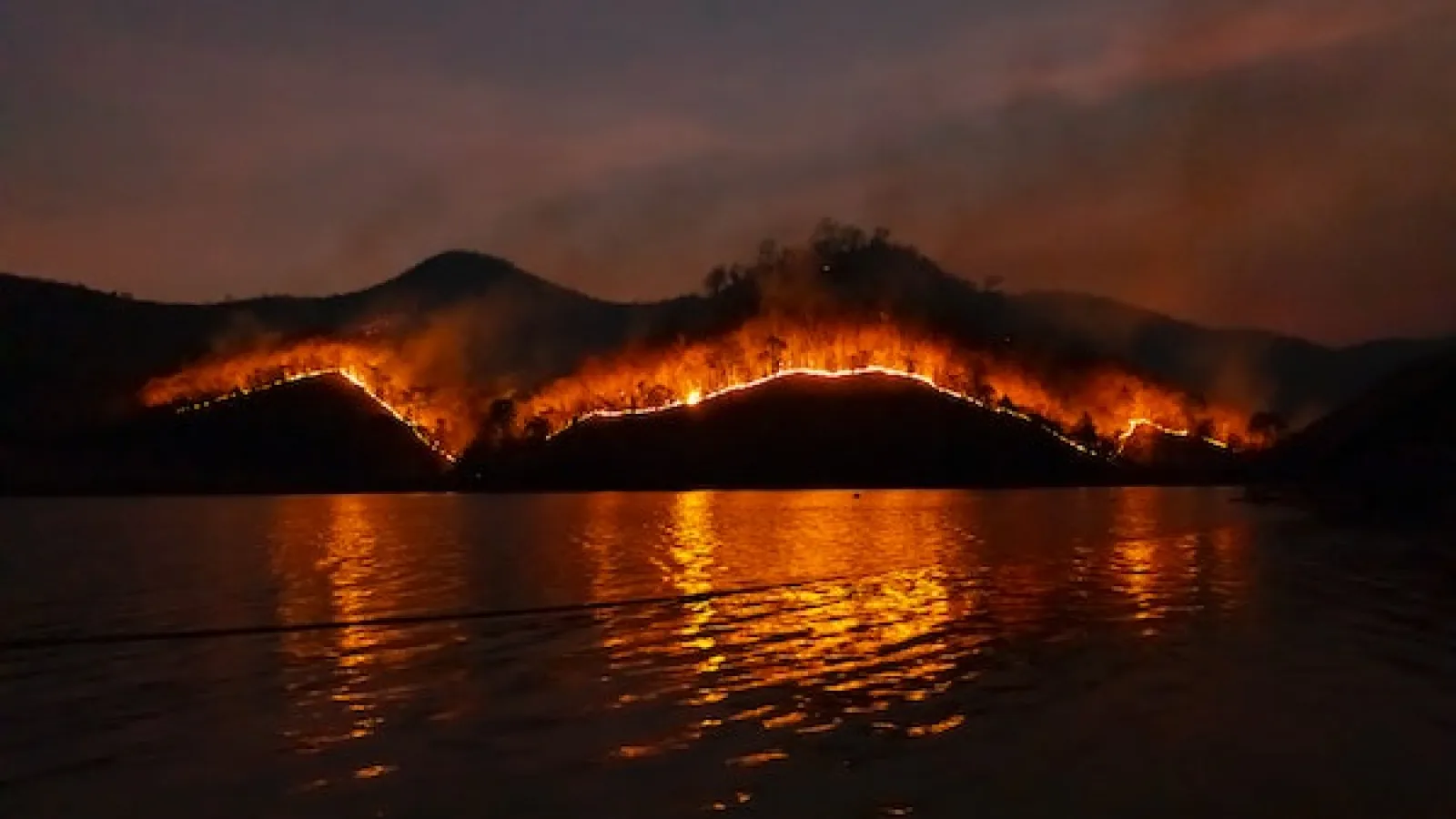 Dusk landscape image of water and some vegetation covered burning hills glowing in a distance with reddish smoke rising from it