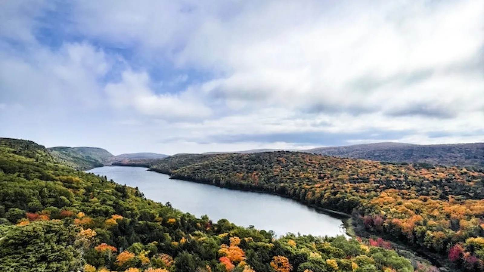 Scenic view from a rock, of a river surrounded by green and orangish leaf trees with white clouds