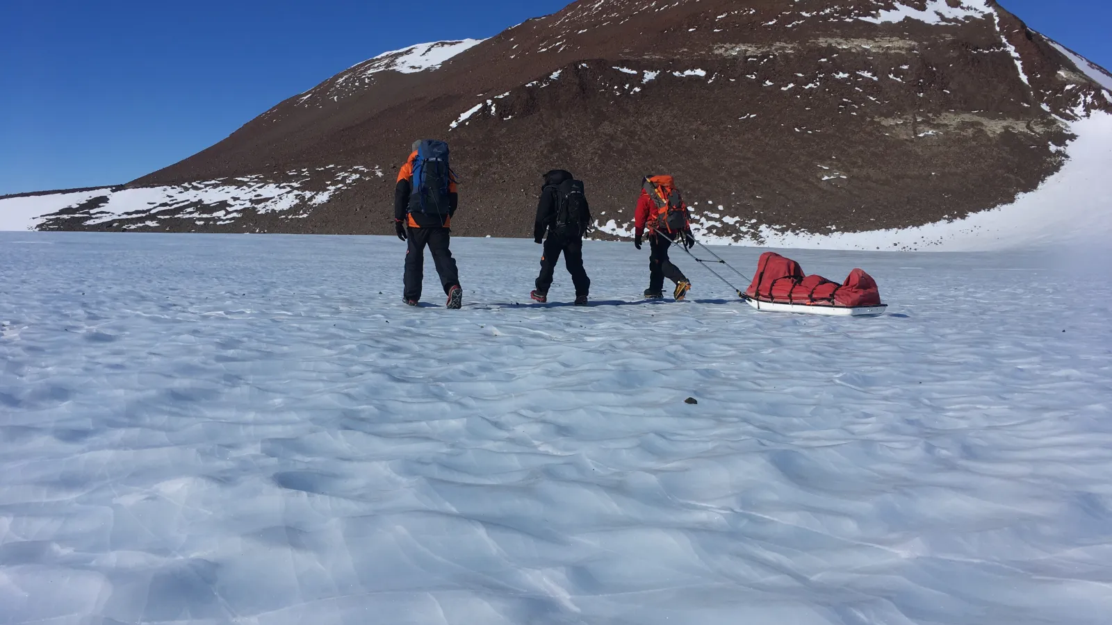Snow covered ground with a  rock mountain and three people walking away from the camera. one pulling a red sled behind him