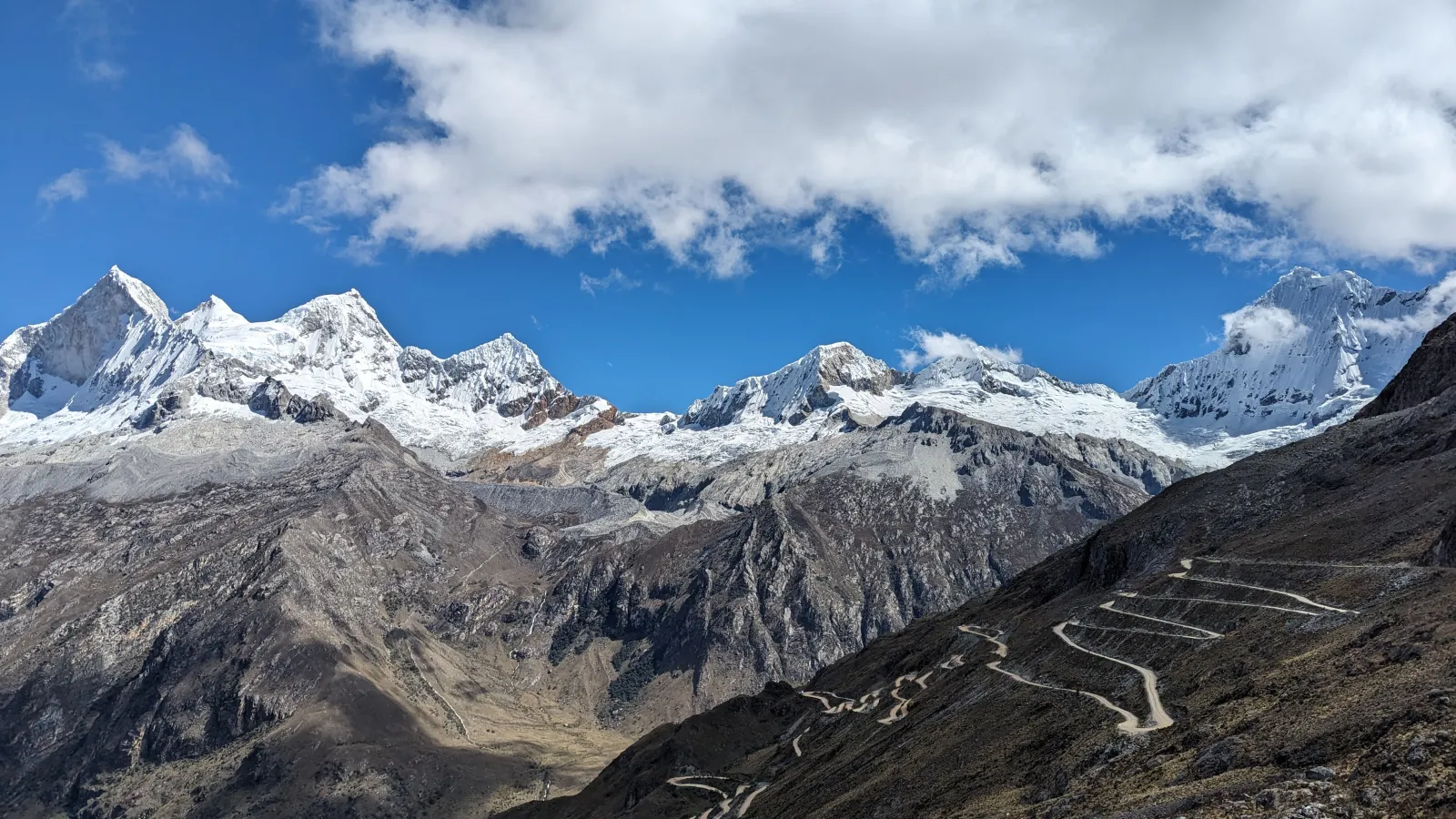 jagged snow capped mountains under blue skies and white clouds. A winding road is carved on the side of one mountain.