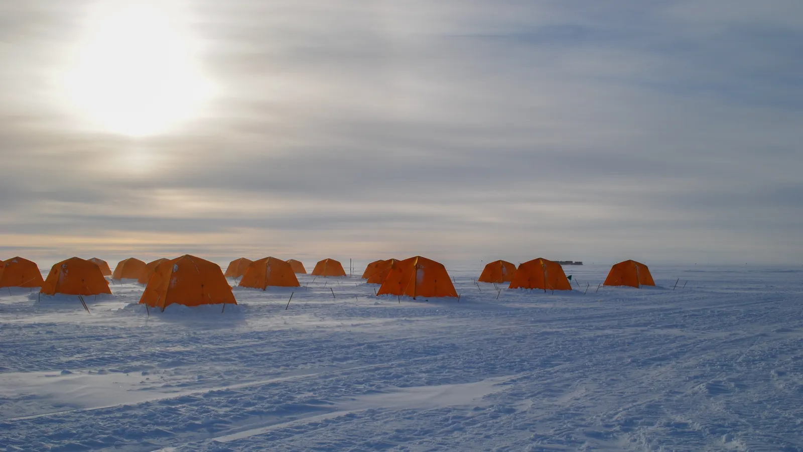 Snowy field with a group of orange tents under cloudy skies with halo of sun.