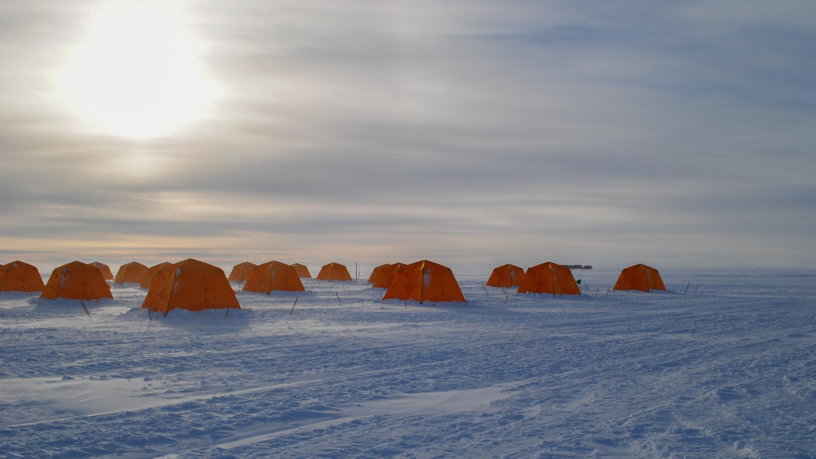 Snowy field with a group of orange tents under cloudy skies with halo of sun.