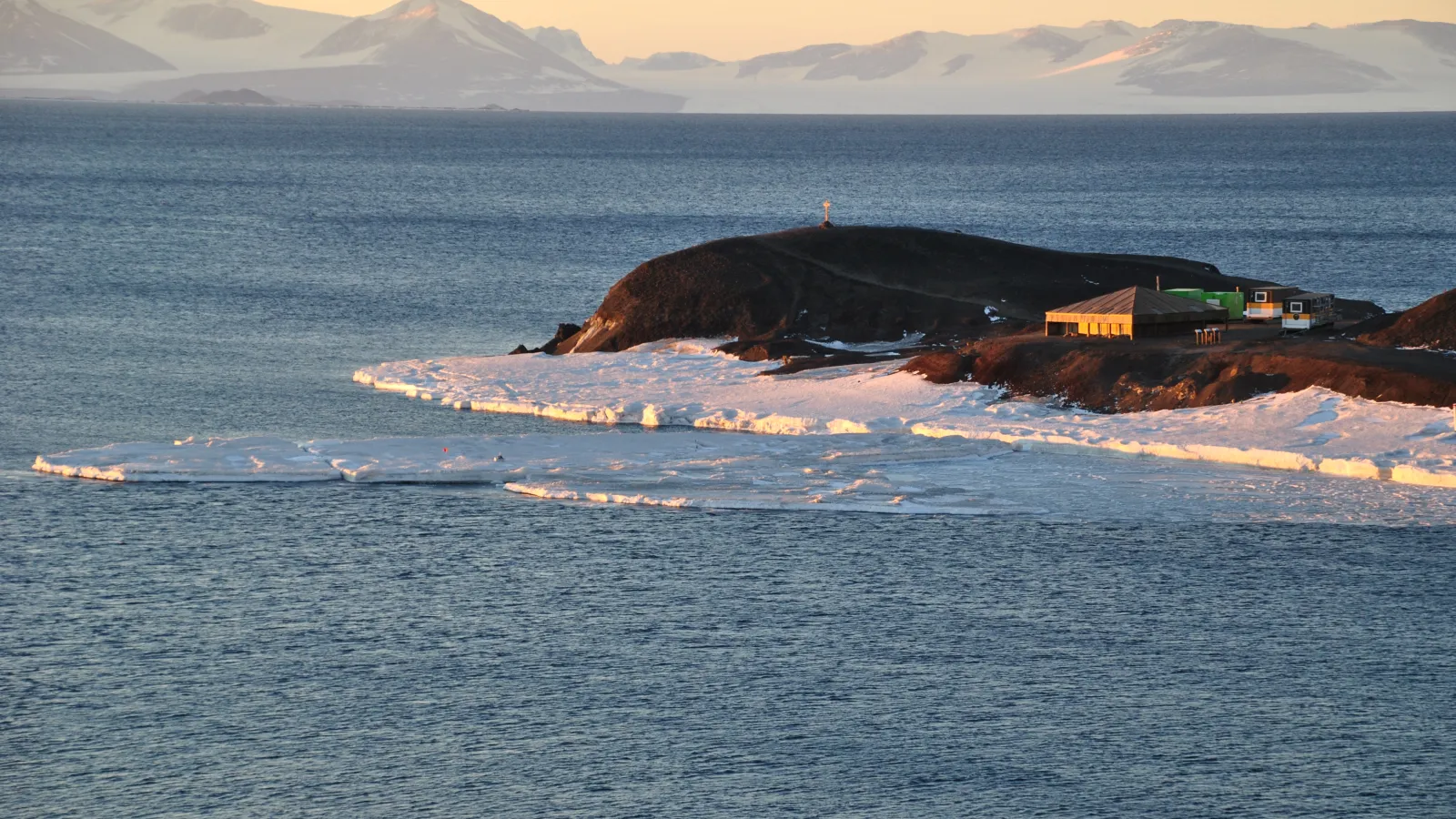 A view of the somewhat frozen lake and a house on a bedrock in a distance with snow covered mountains  in the background under cream colored sky,