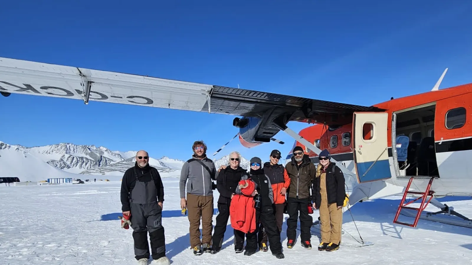 Several people posing next to a plane on a sunny day with blue skies in winter gear with snow covered mountains in the background and snow on the ground.
