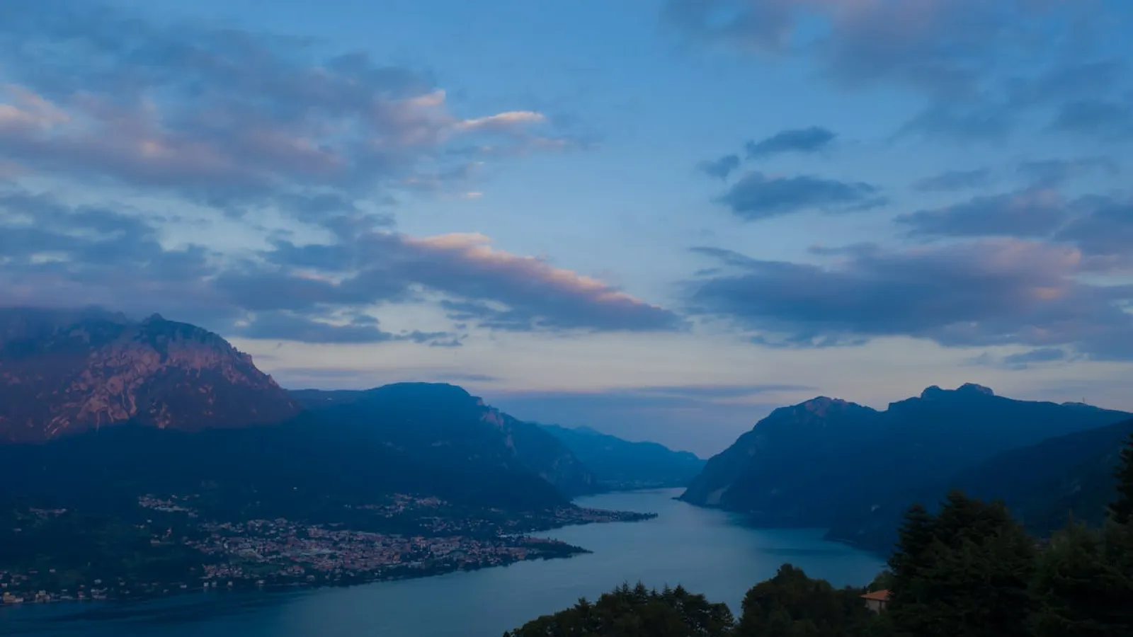 Mountains covered with trees surrounding a river with a city on one side during day break with some clouds.