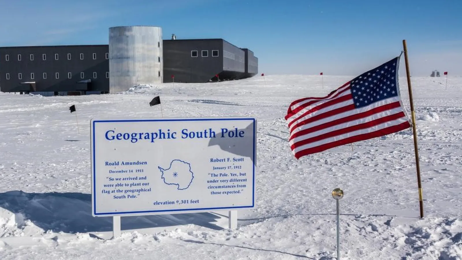 Snowy ground with a sign and flag planted in the ground and a dark building in the background under blue skies.