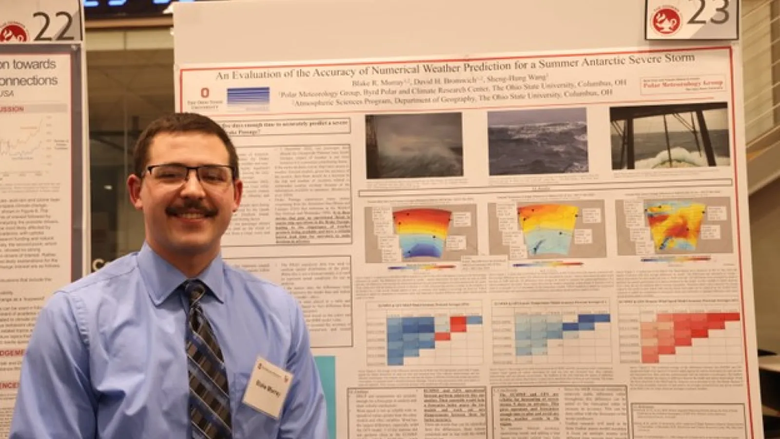 A man in blue dress shirt with a tie and glasses standing in front of a poster.