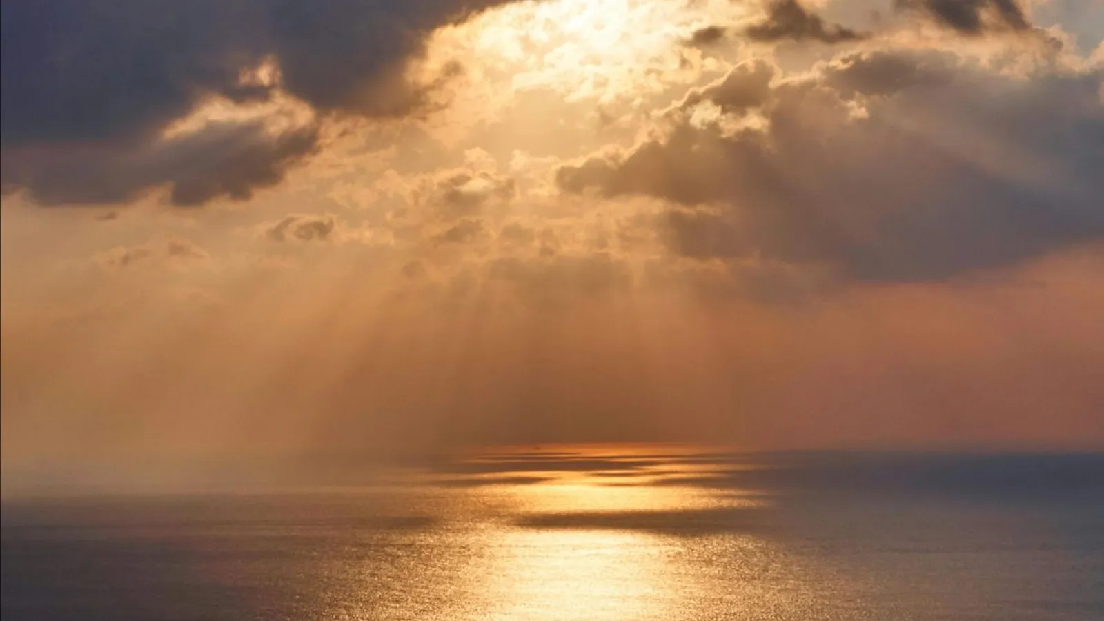 Vast, calm Ocean as far as the eye can see under dark clouds with rays of orange sunlight shining down.