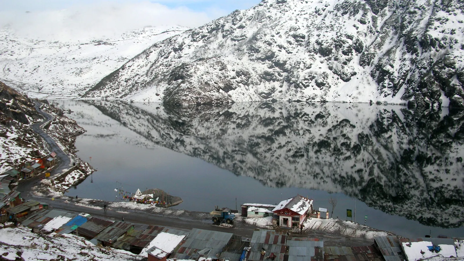 A lake surrounded by snow covered mountains with tin roof huts and a road along its perimeter in the foreground under blue skies and white clouds..