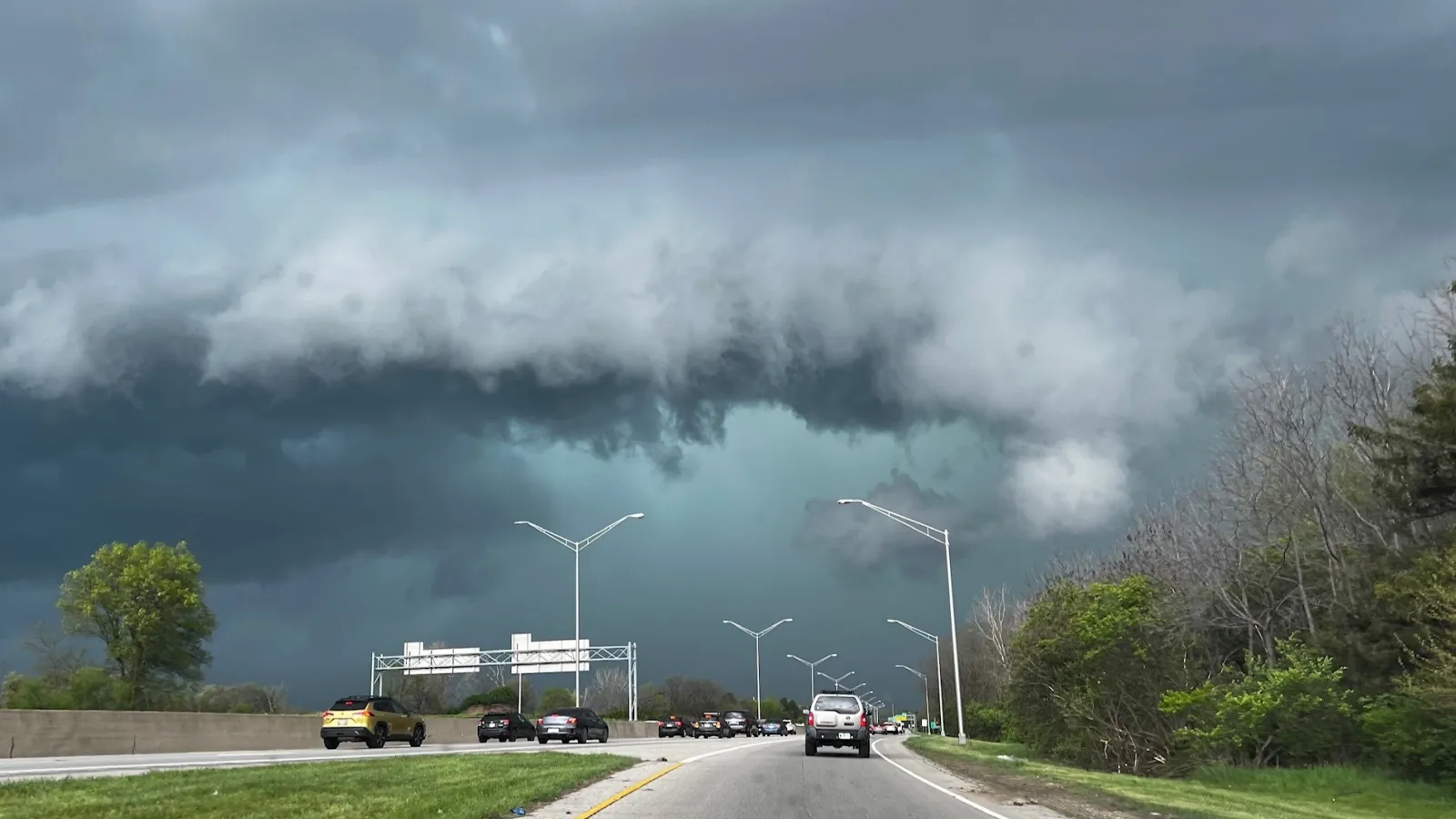 Freeway with cars in different lanes, streetlamps and trees lining the roadside and cloudy skies above