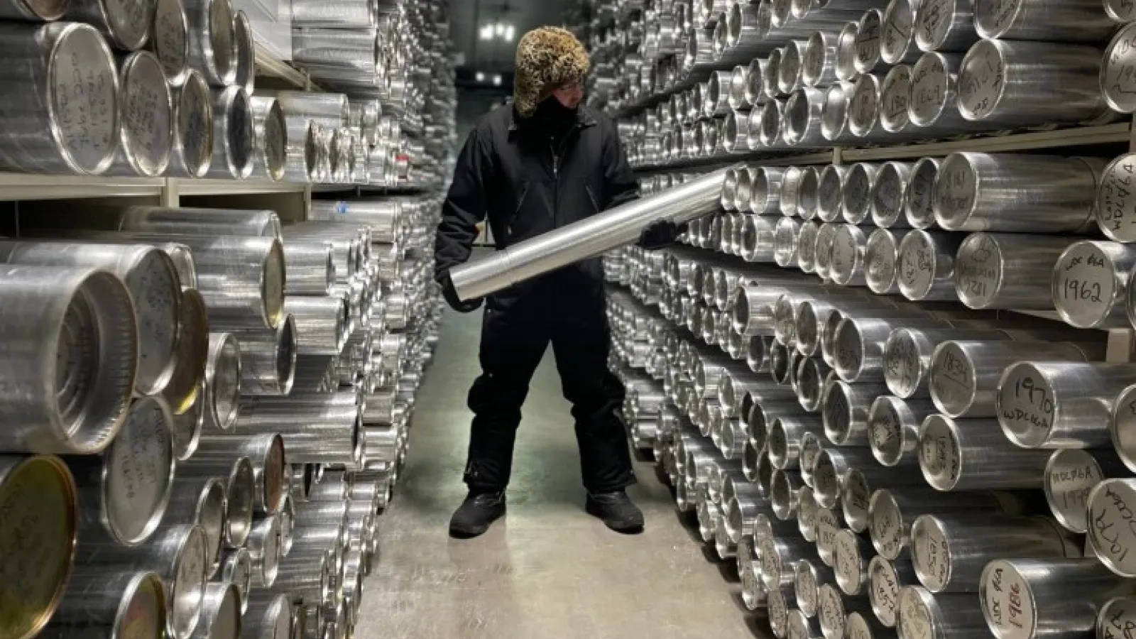 A man wearing dark color winter gear, hat and gloves, standing in a room. He is holding a long silver cylinder in both hands surrounded on both sides with shelves of silver cylinders.