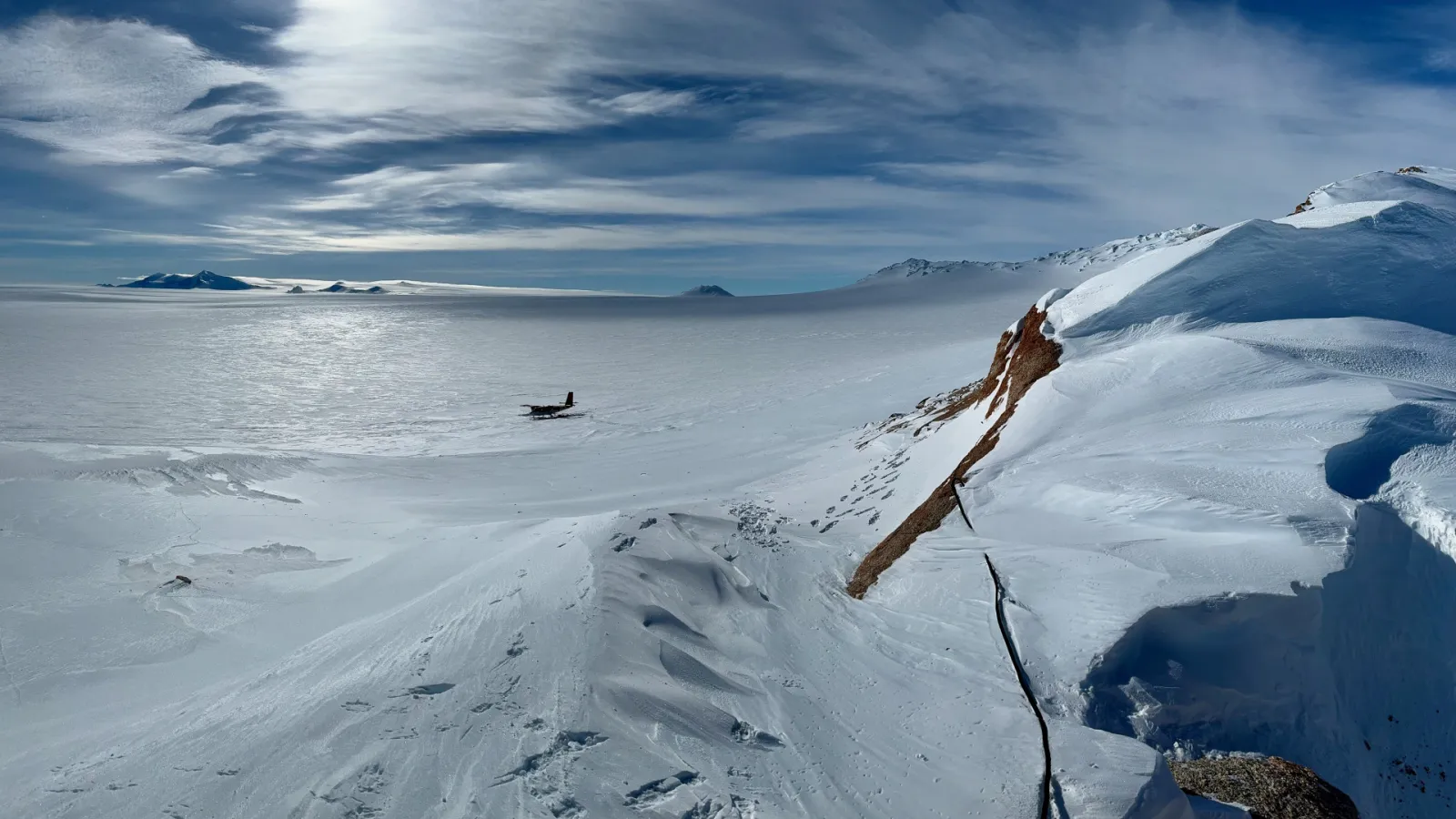 A vast area covered in snow and ice with mountains and a small plane in the snow at a distance.