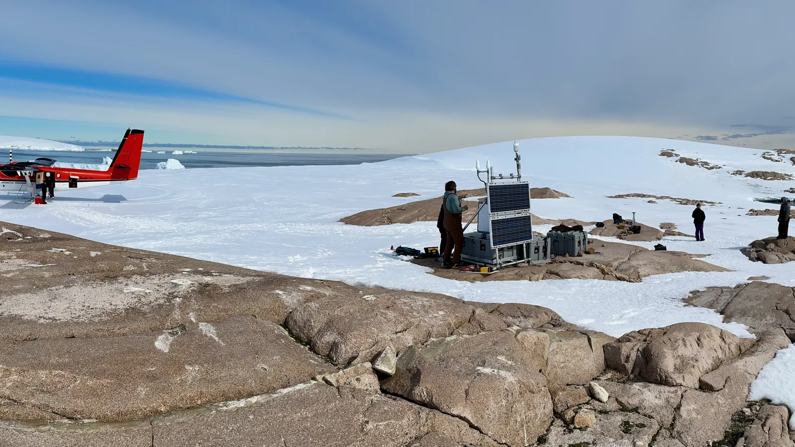A small plane in a snow-covered ground with the background of sea with icebergs in it and people and a bedrock in the foreground working on equipment.