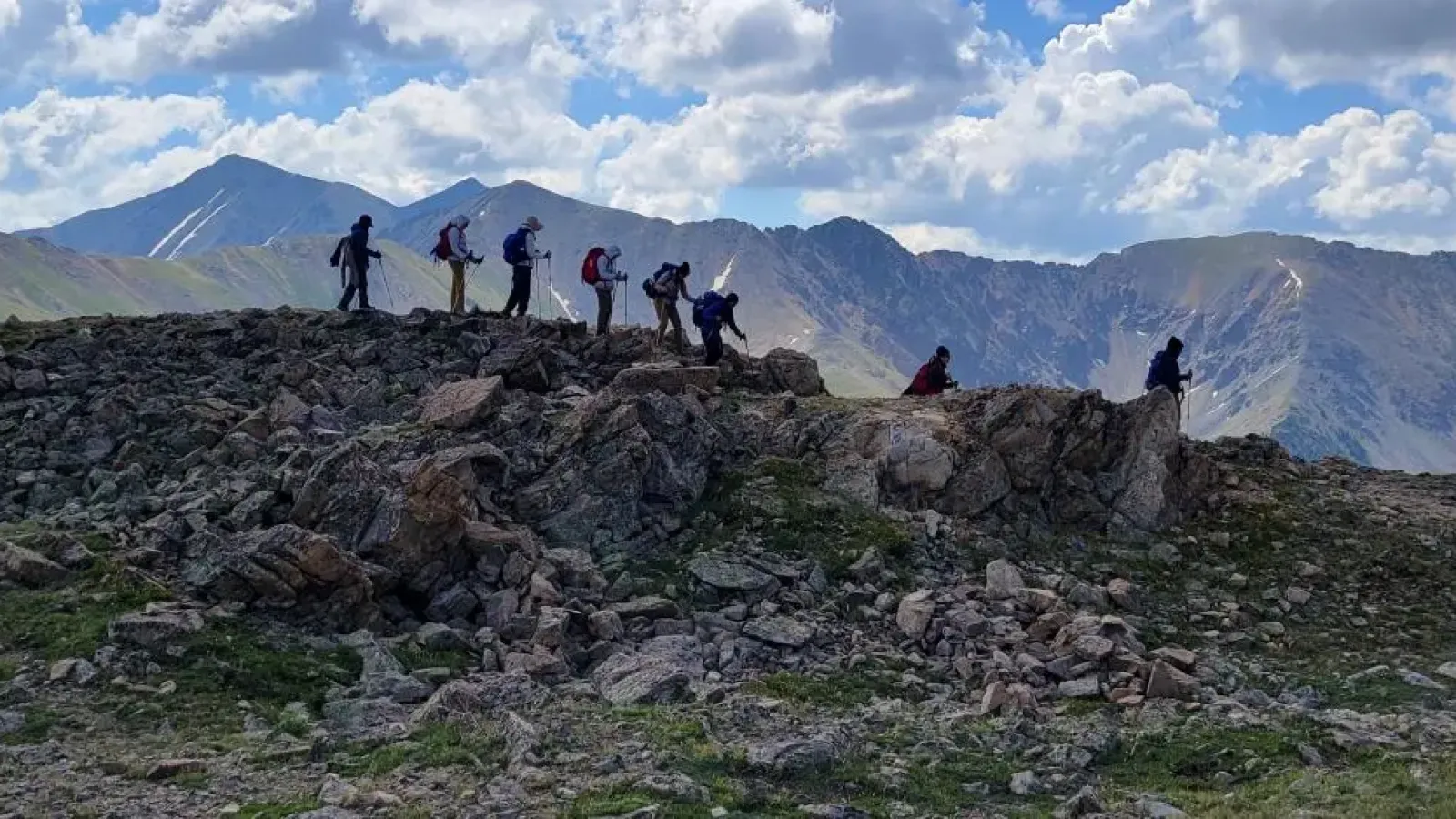 A group of eight with backpacks and walking gear walking along a rocky path with a backdrop of mountains, under blue skies and white clouds.