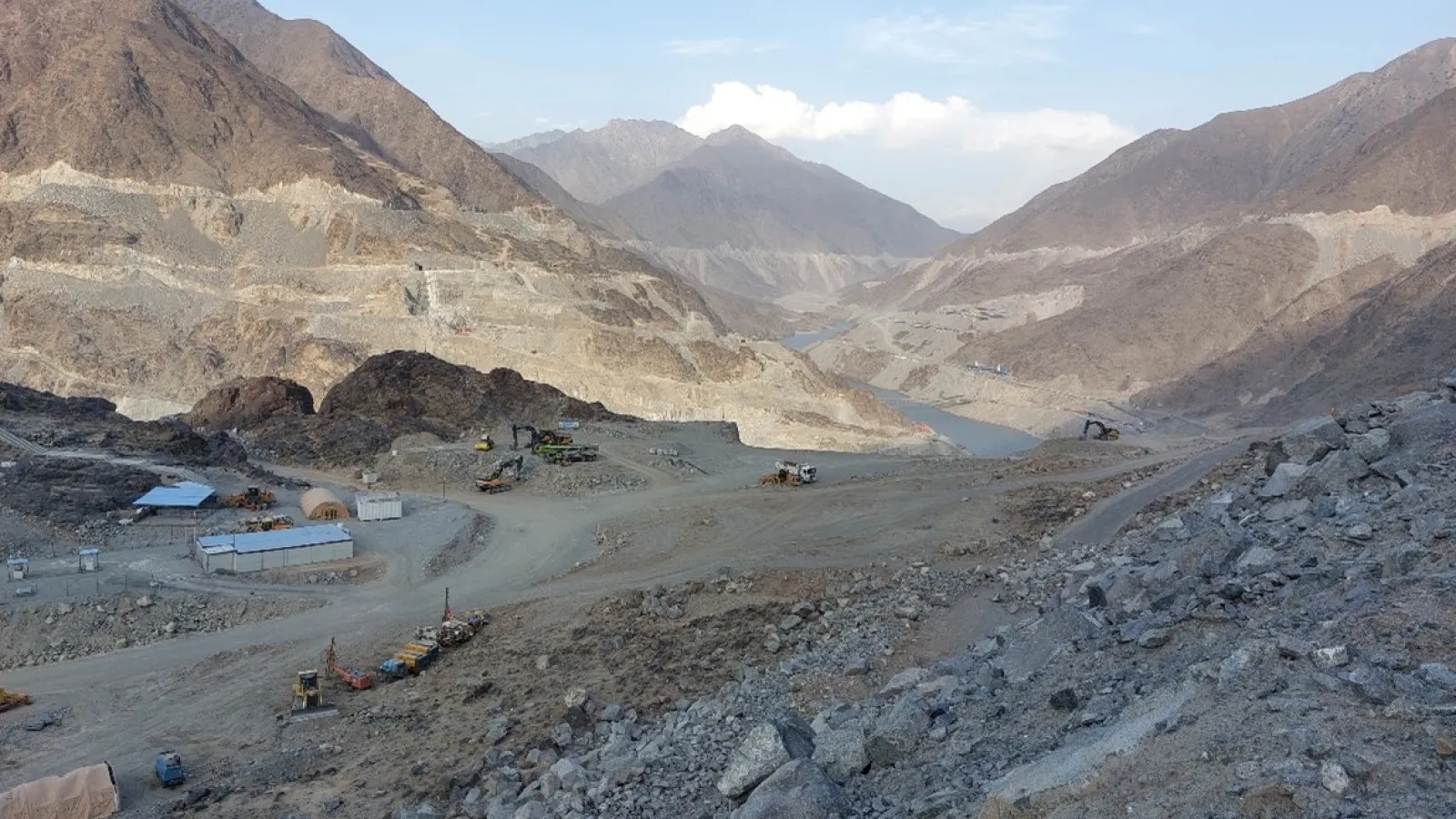 a low lying river in a rocky-dirt terrain in a valley surrounded by brown mountains with some structures, trucks and machinery under blue skies and white clouds.