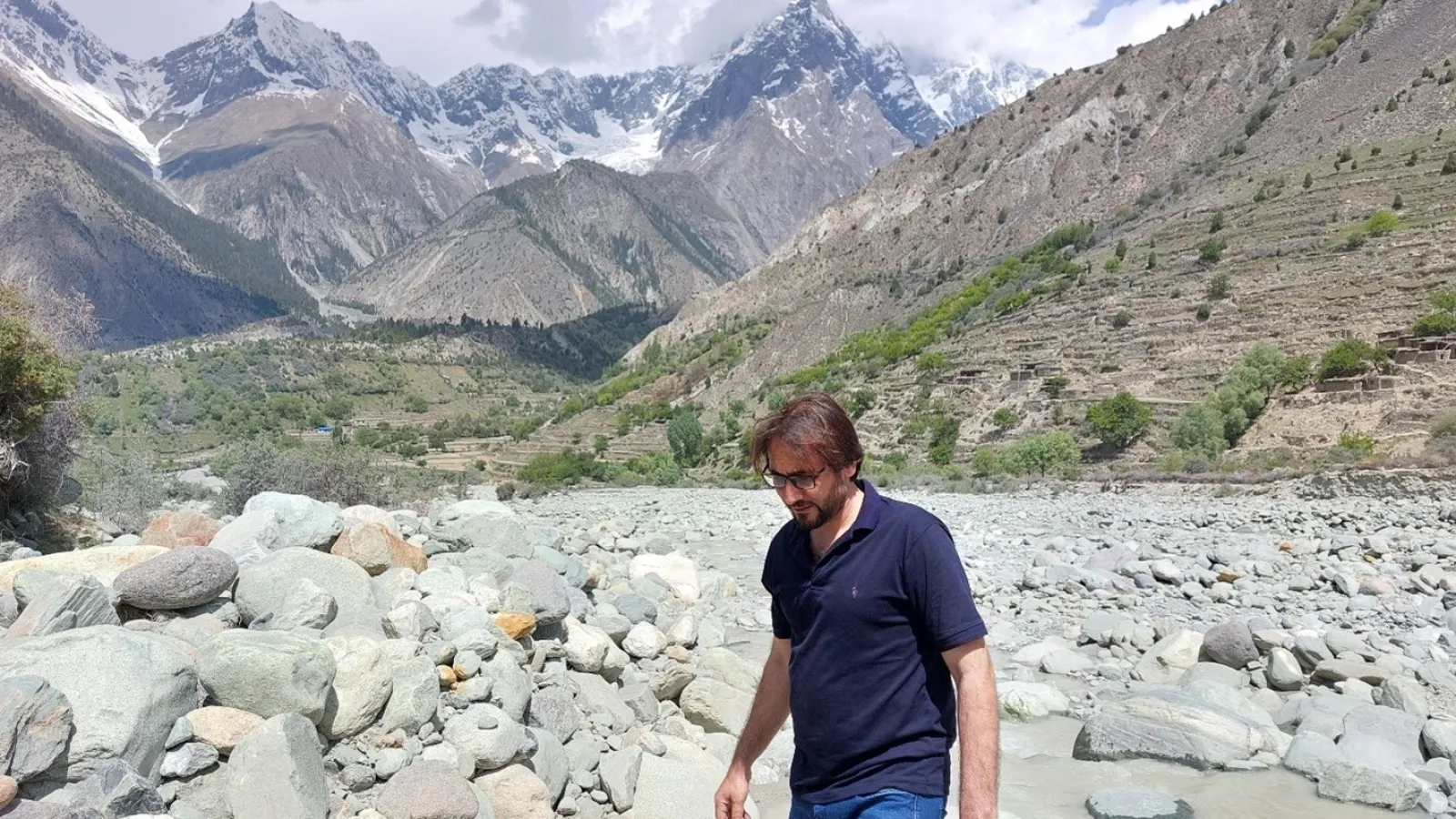 A man holding a green-covered notebook walking in a rocky river bed with a little stream flowing behind him and sparse tree covered hills and mountains in the background. 