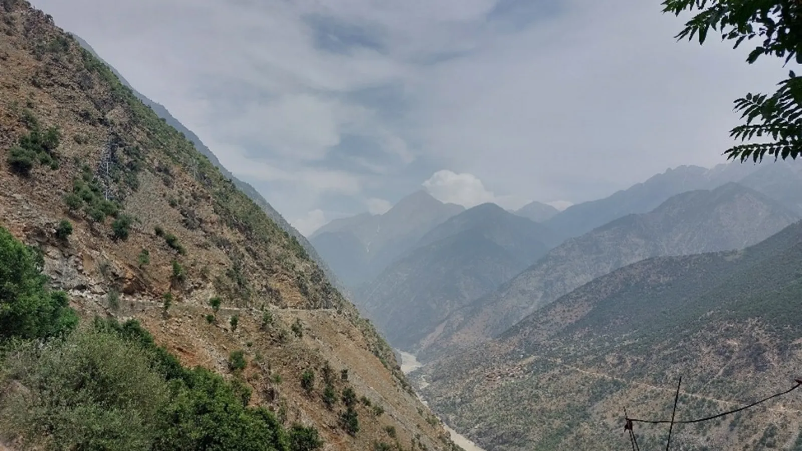 A dirt Mountain range covered sparsely with trees and a river flowing with light brown water below.