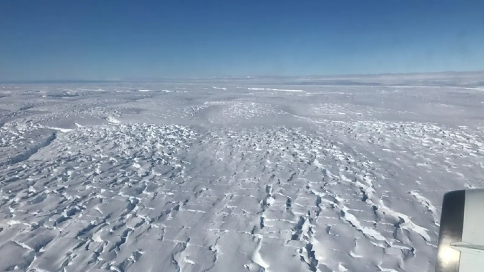 Vast area covered in snow and ice under blue skies with a jet engine visible in the bottom right corner. 